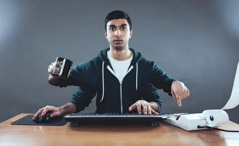 Homme à son bureau doté de quatre bras, tenant à la fois une tasse de café, la souris et le clavier : métaphore des tâches marketing menées en parallèle