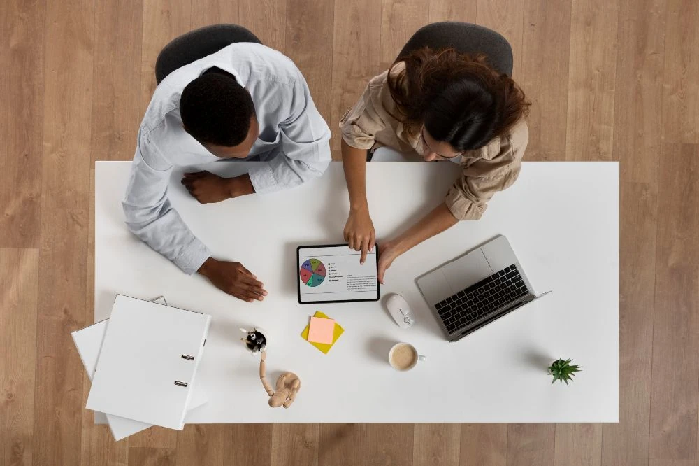 Vue de dessus d’un bureau blanc : deux personnes consultent ensemble un tableau de bord de données clients sur une tablette, à côté d’un ordinateur portable et d’un carnet