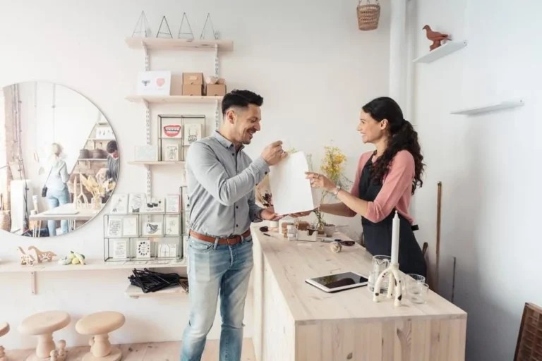 Dans une boutique de décoration lumineuse, une commerçante souriante remet un sac en papier à un client au comptoir