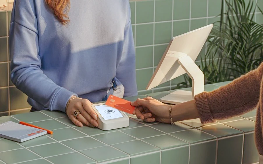 Au comptoir carrelé de vert d’une boutique, une cliente approche sa carte bancaire d’un terminal de paiement tenu par la commerçante