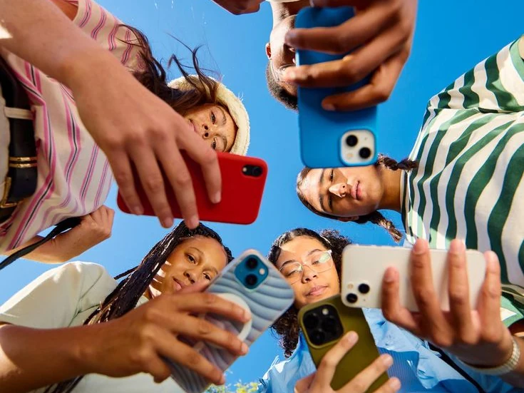 Vue en contre-plongée d’un groupe de jeunes réunis en cercle, chacun tenant son téléphone, sous un ciel bleu