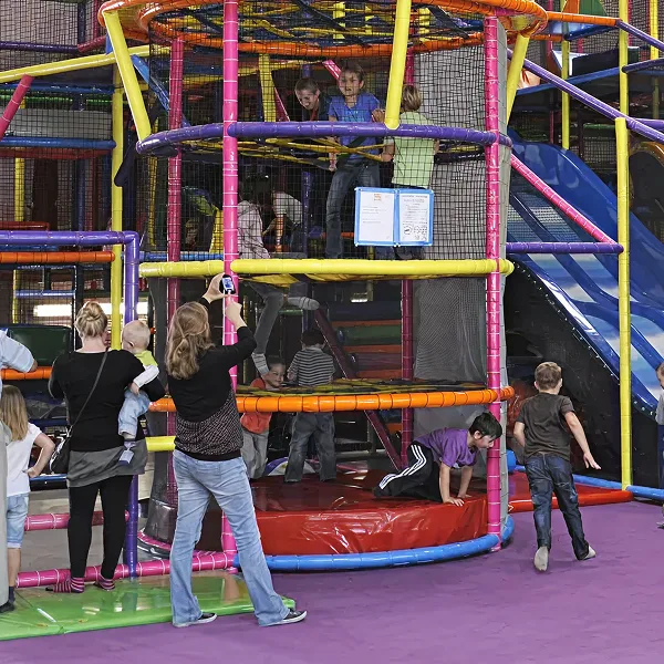 Enfants jouant dans une structure de jeux colorée avec toboggan en intérieur, tandis que des adultes les surveillent.